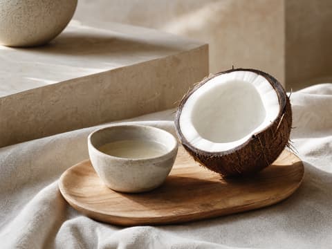 A ceramic bowl of coconut oil beside half a coconut on a wooden tray